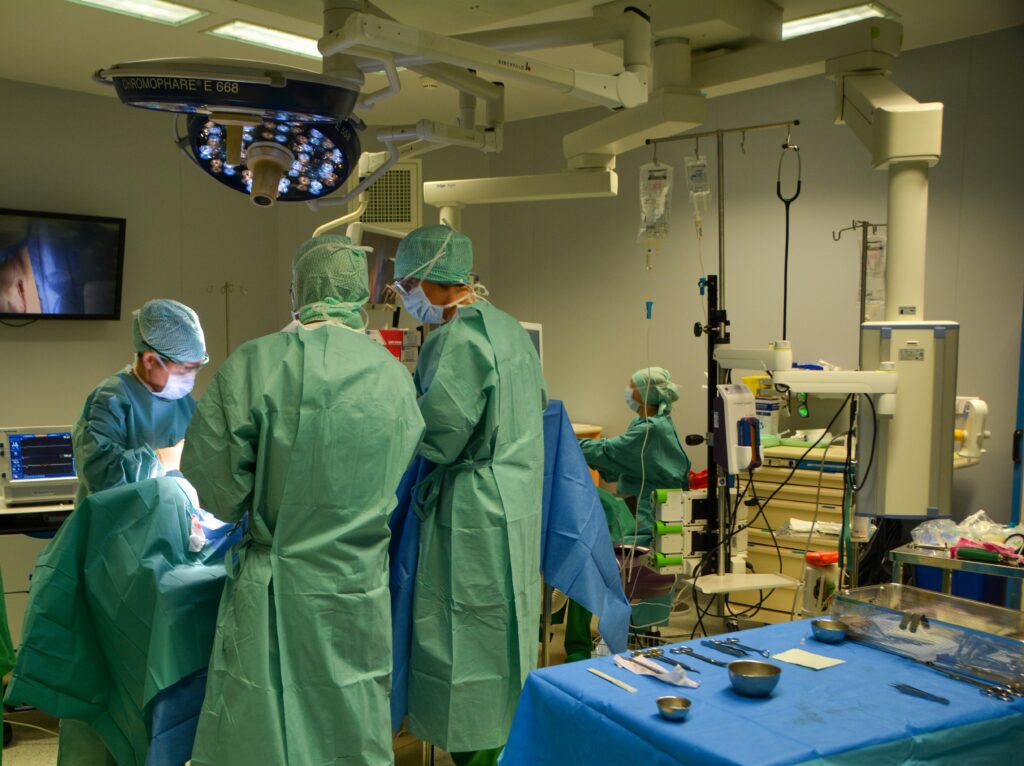 Surgeons in green gowns and masks performing an operation in a sterile operating room with overhead surgical lights and monitors.